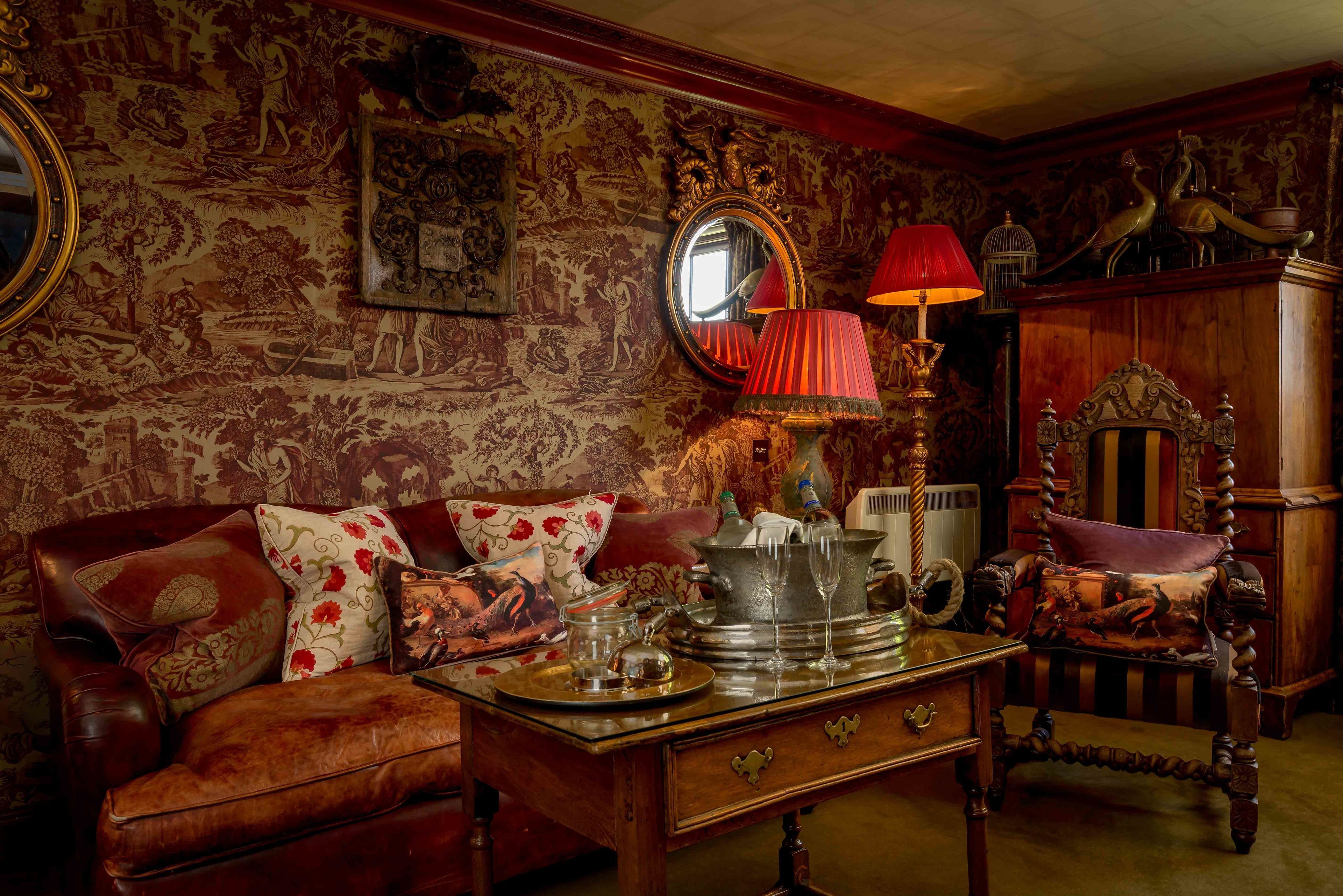 Una habitación de estilo antiguo con papel tapiz ornamentado, una mesa de madera con lámpara, un espejo y un sofá mullido.
