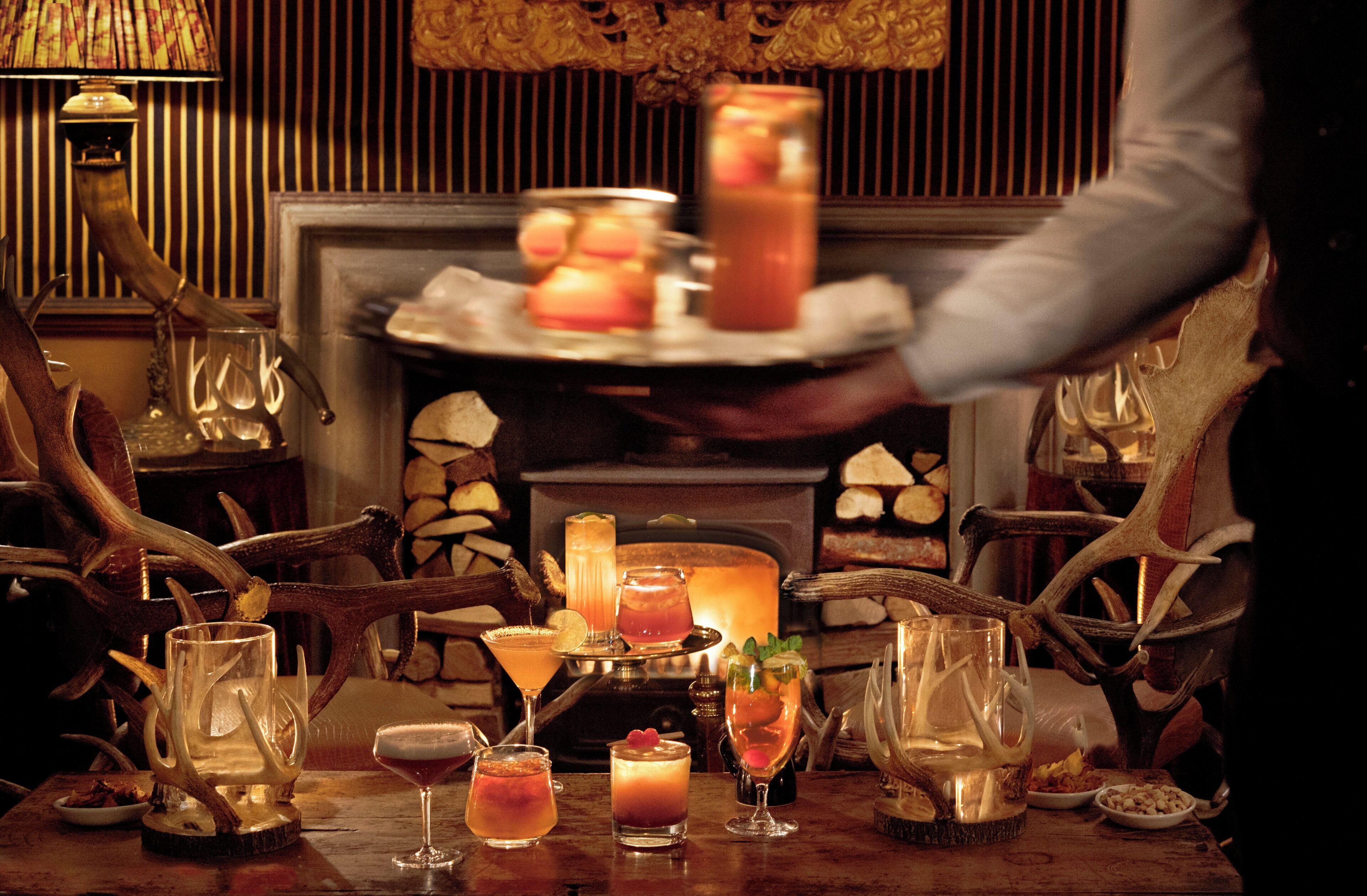 A table set with various glasses and a tray of food, a fireplace in the background, and antlers as decor.