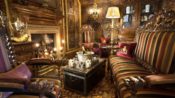 Una habitación ricamente decorada con muebles ornamentados, una chimenea y una variedad de objetos decorativos, incluyendo un baúl, lámparas y un candelabro.