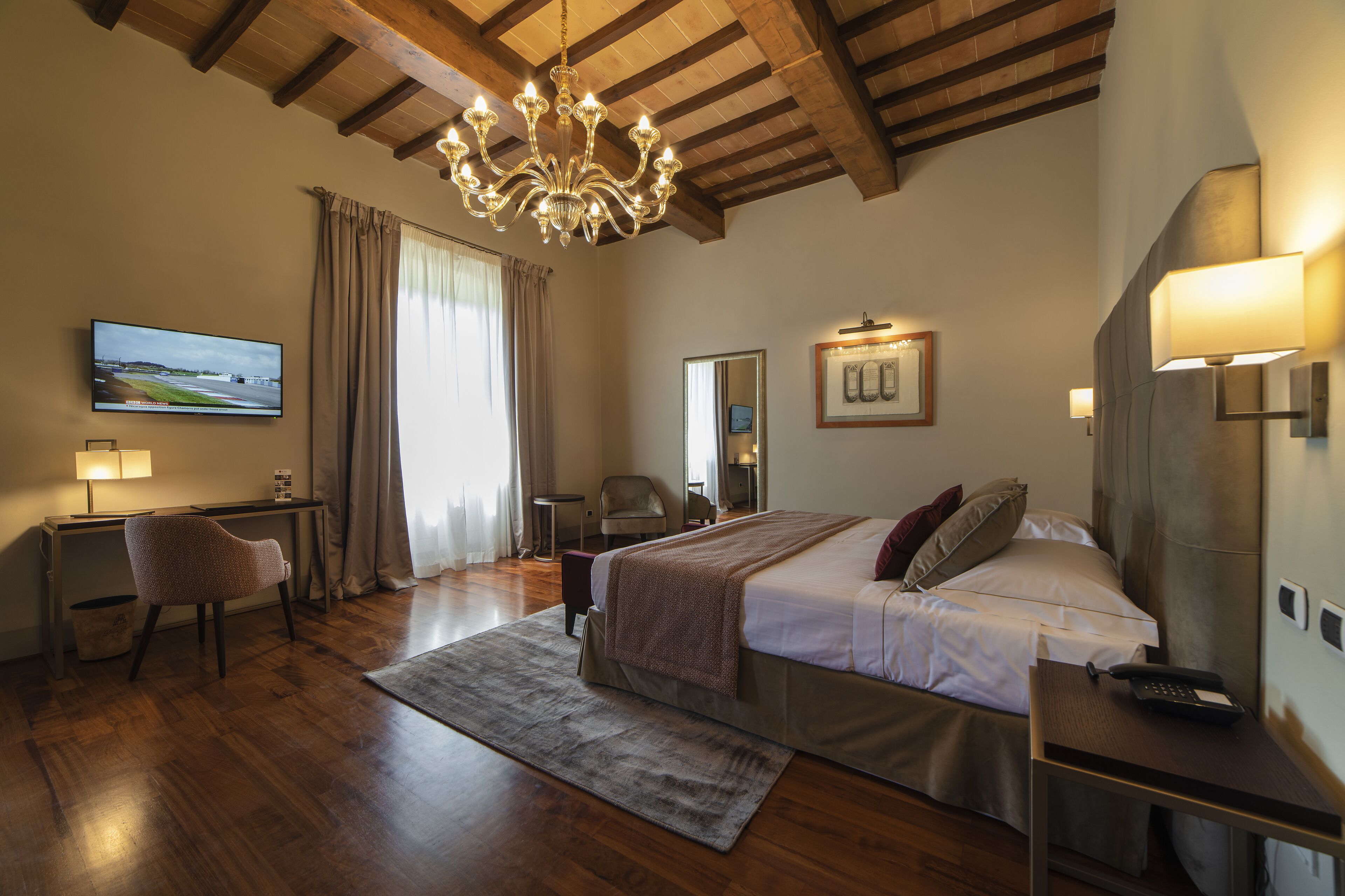 Una camera d'albergo con soffitto in legno, un letto con coperta, una scrivania con lampada, una sedia, una televisione e una finestra con tende.