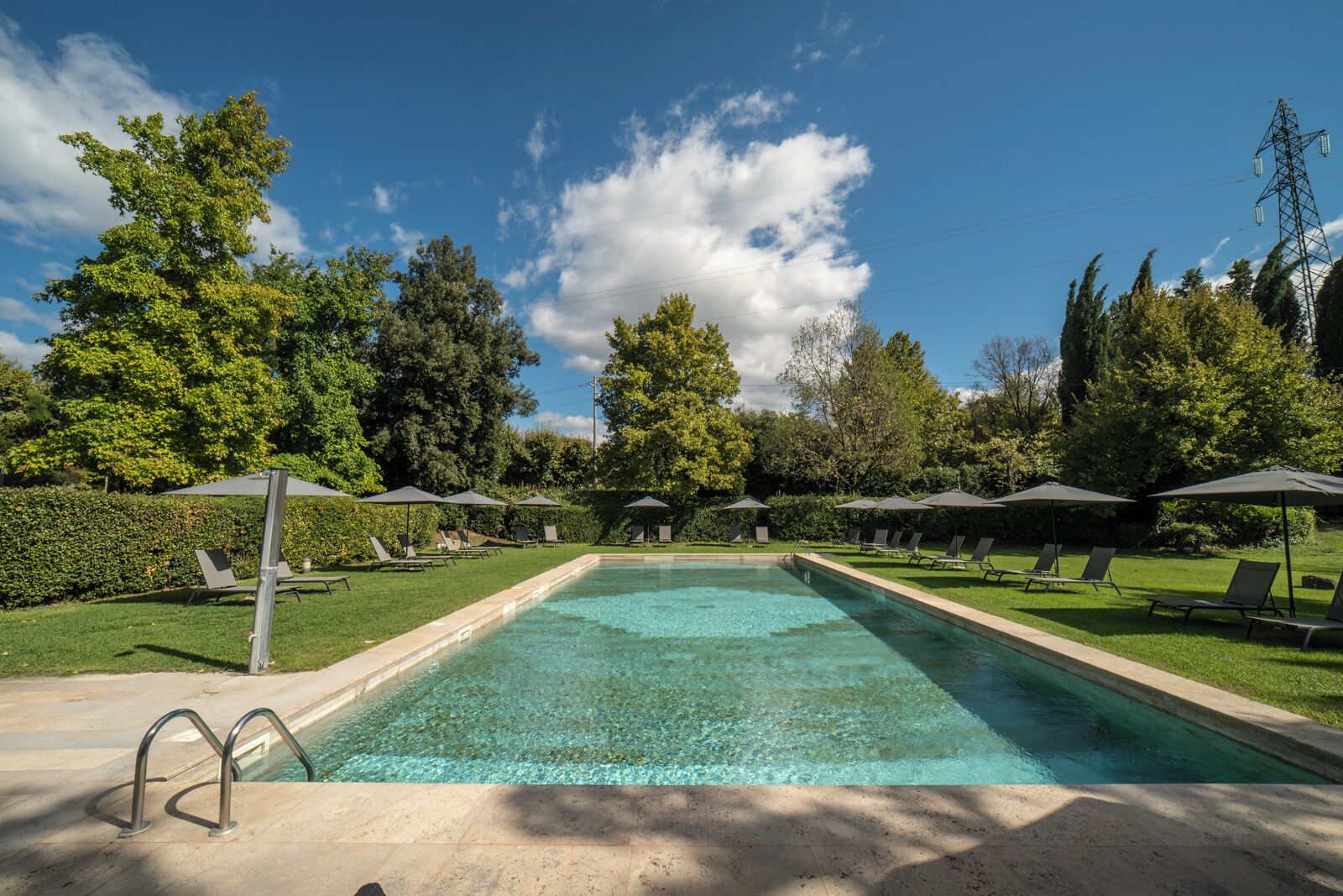 Piscina rectangular con agua cristalina, rodeada de sillones de descanso y sombrillas, con árboles y un cielo despejado de fondo.