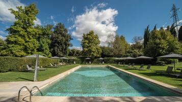 Piscina rectangular con agua cristalina, rodeada de sillones de descanso y sombrillas, con árboles y un cielo despejado de fondo.