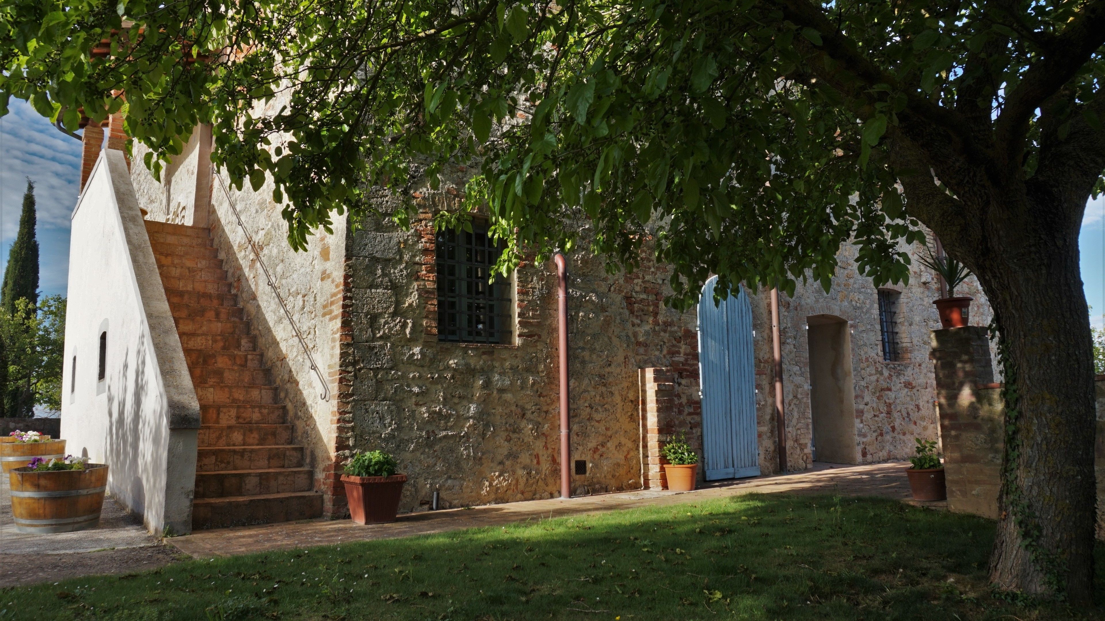 A rustic stone building with a staircase, wooden barrels, and potted plants.