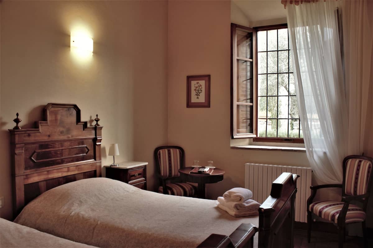 Une chambre d’hôtel comprenant un lit, deux chaises, une petite table, une table de chevet, une lampe et une fenêtre avec des rideaux.