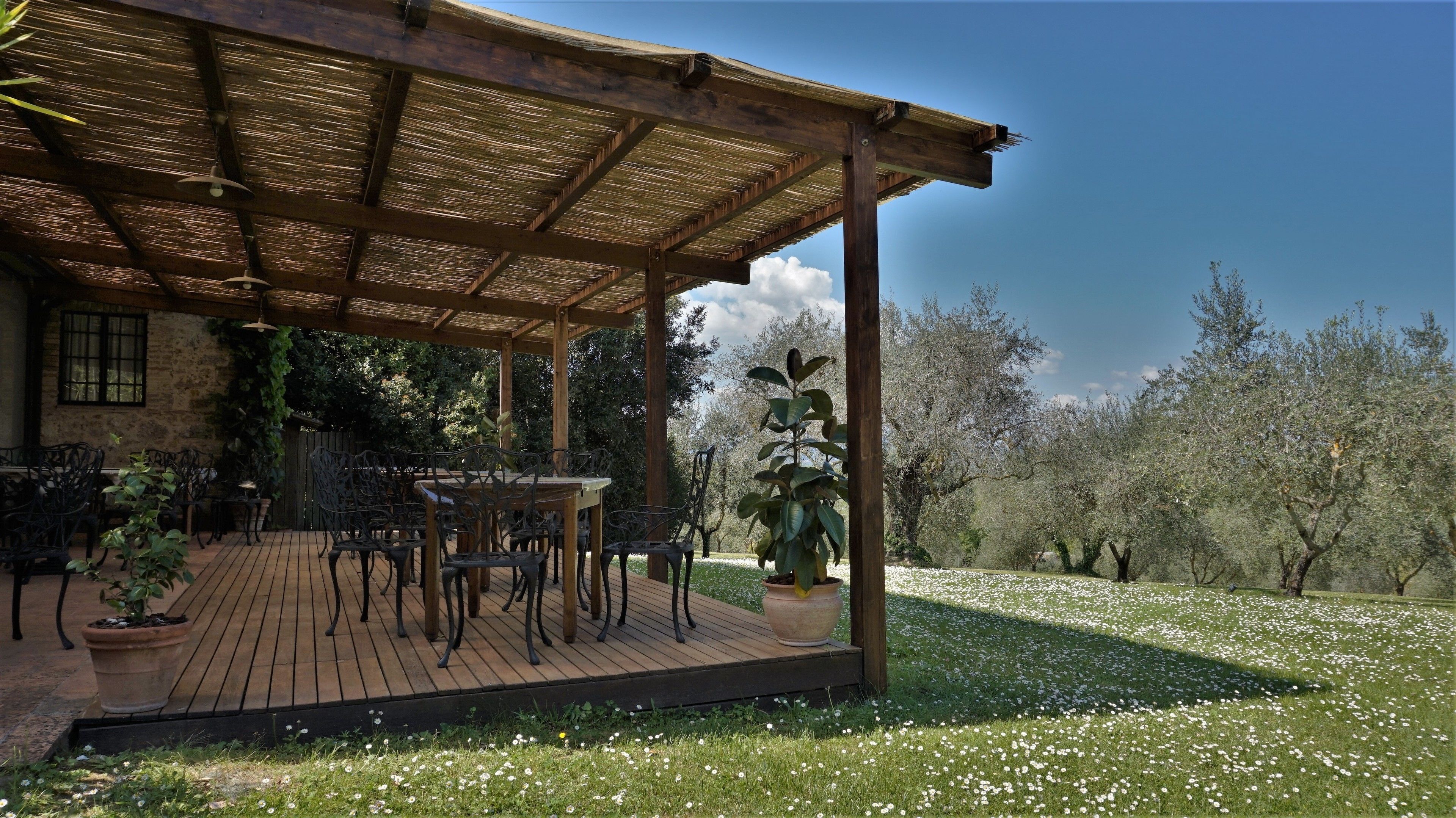 Eine überdachte Terrasse mit Holzboden und Metallstühlen, umgeben von einem Garten mit weißen Blumen und Bäumen.