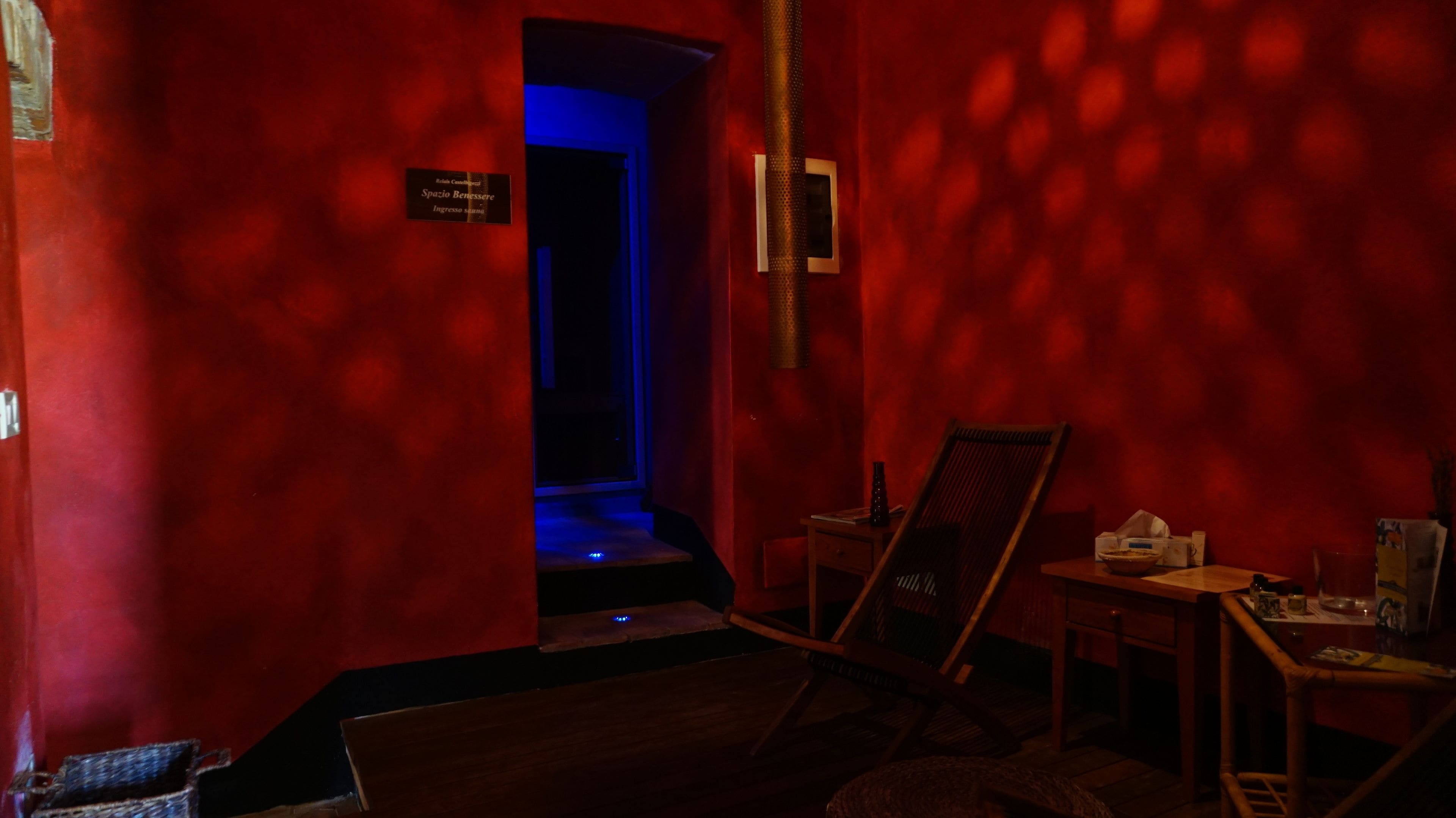 Une pièce faiblement éclairée aux murs rouges, avec une chaise en bois, une petite table sur laquelle se trouvent divers objets, et une pancarte indiquant : « Spazio Benessere, entrée de la salle ».