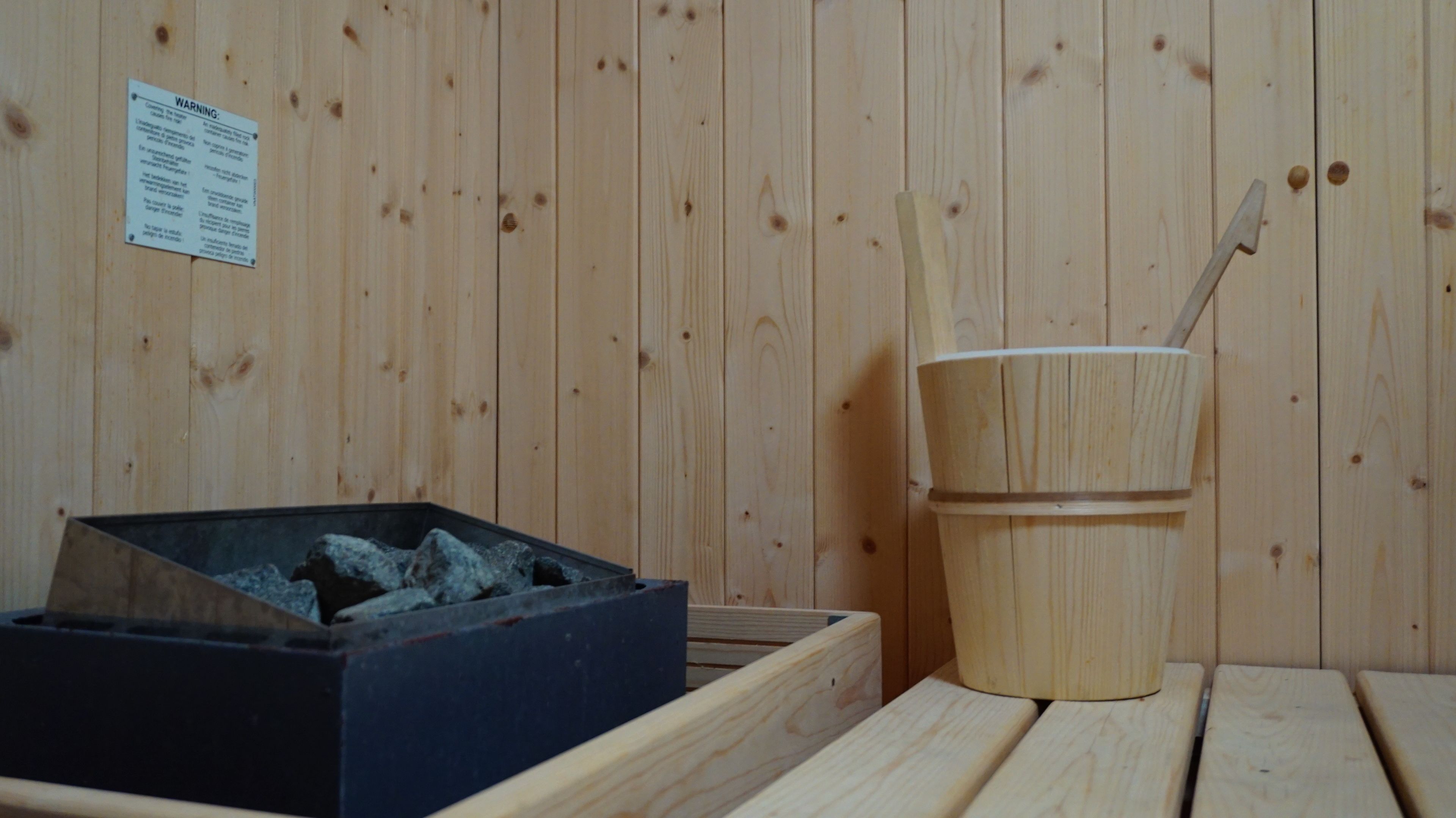 Un cuarto de sauna con paredes de madera, un banco de madera, un cubo con una cuchara y una caja de piedras.