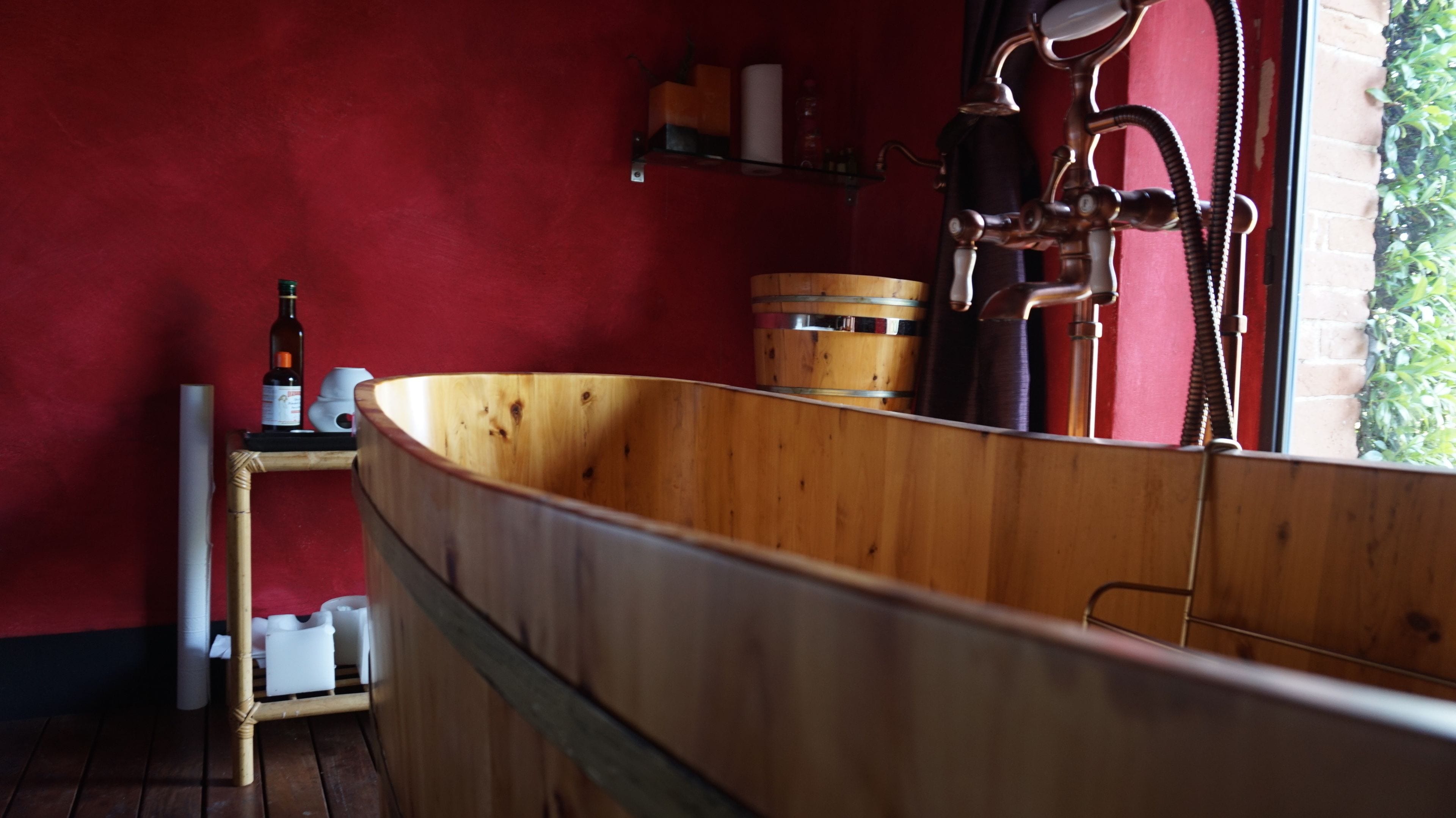 A wooden bathtub in a room with a red wall, a small shelf with bottles, and a window with a view of greenery.