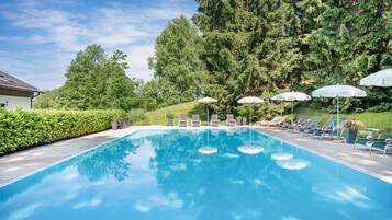 Ein Schwimmbecken mit Liegestühlen und Sonnenschirmen, umgeben von Bäumen und einer Hecke.