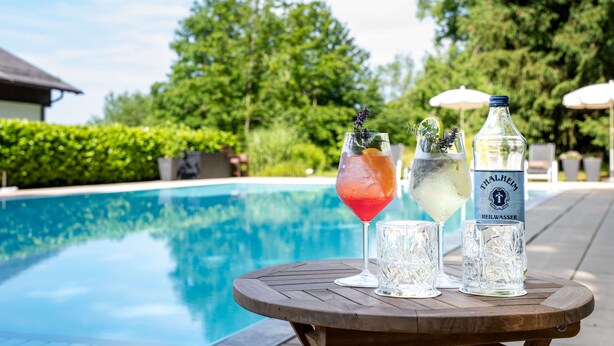A poolside table with drinks and a bottle, surrounded by lush greenery and a clear pool. - Romantik Hotel die Gersberg Alm mit Panorama über Salzburg und Outdoorpool ab Mai (Salzburg)