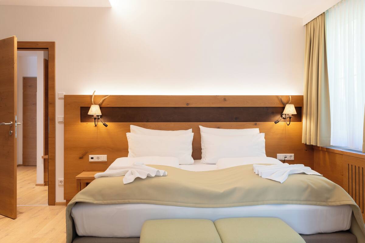 A hotel room with a wooden headboard, two bedside lamps, a bed with white linens, and a window with curtains.
