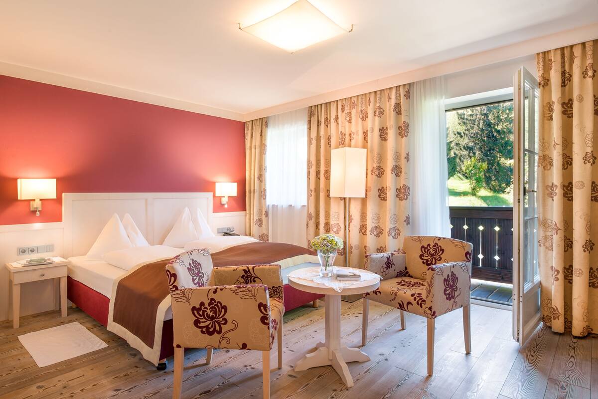 A hotel room with a bed, two chairs, a small table, and a view of the outdoors.