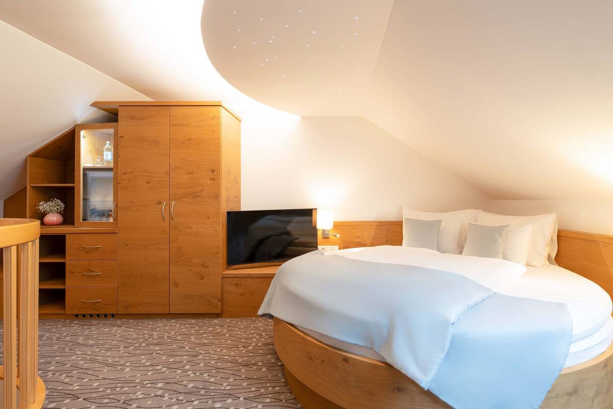A bedroom with a wooden wardrobe, a television, a bed with white bedding, and a patterned carpet.