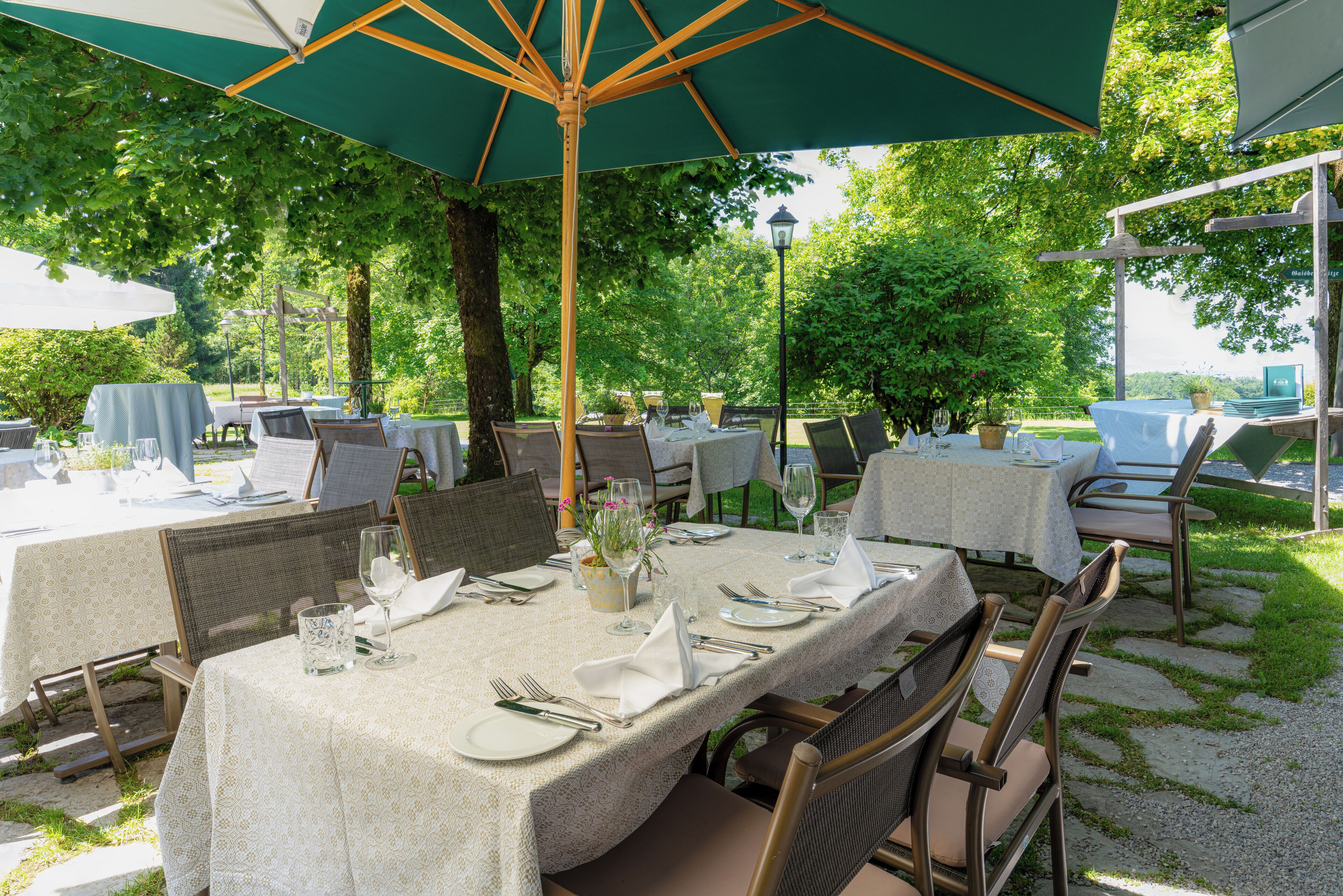 An outdoor dining area with tables set for a meal, surrounded by trees and covered by umbrellas.