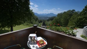 A balcony with a table set for two, featuring a bottle of champagne, two glasses, and a fruit plate. The balcony overlooks a scenic view of mountains and a garden with red flowers. - Romantik Hotel die Gersberg Alm mit Panorama über Salzburg und Outdoorpool ab Mai (Salzburg)
