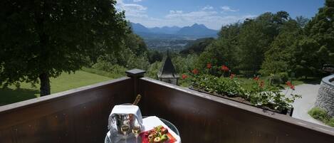 A balcony with a table set for two, featuring a bottle of champagne, two glasses, and a fruit plate. The balcony overlooks a scenic view of mountains and a garden with red flowers.