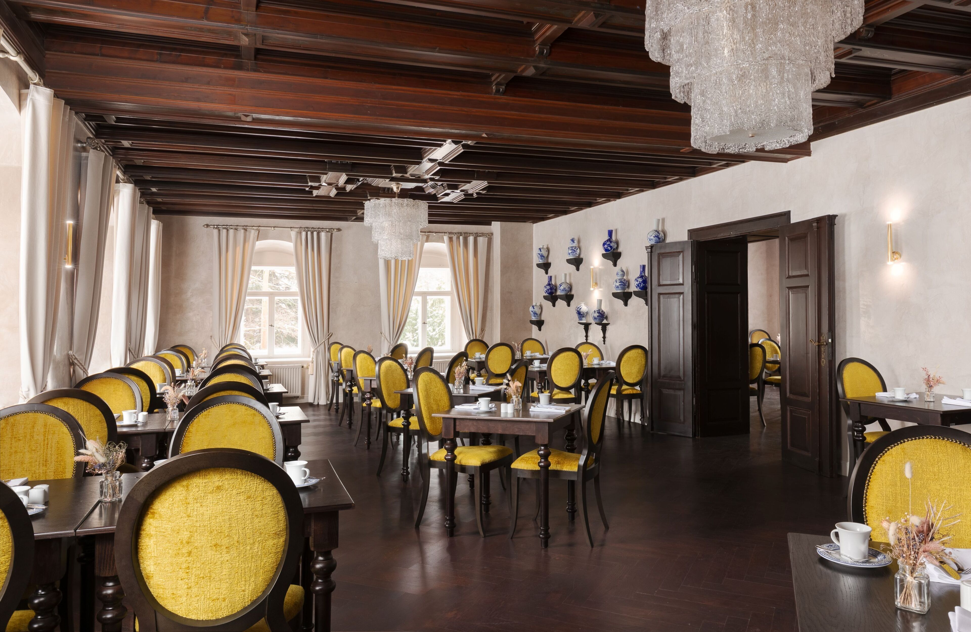 A formal dining room with wooden tables and chairs, chandeliers, and decorative vases on the walls.