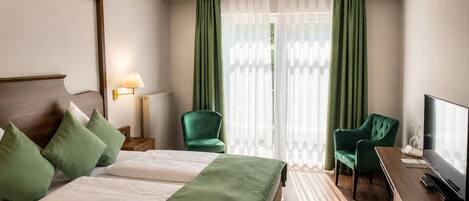 Habitación de hotel con una cama, dos sillones verdes, un escritorio con televisor y una ventana con cortinas traslúcidas.