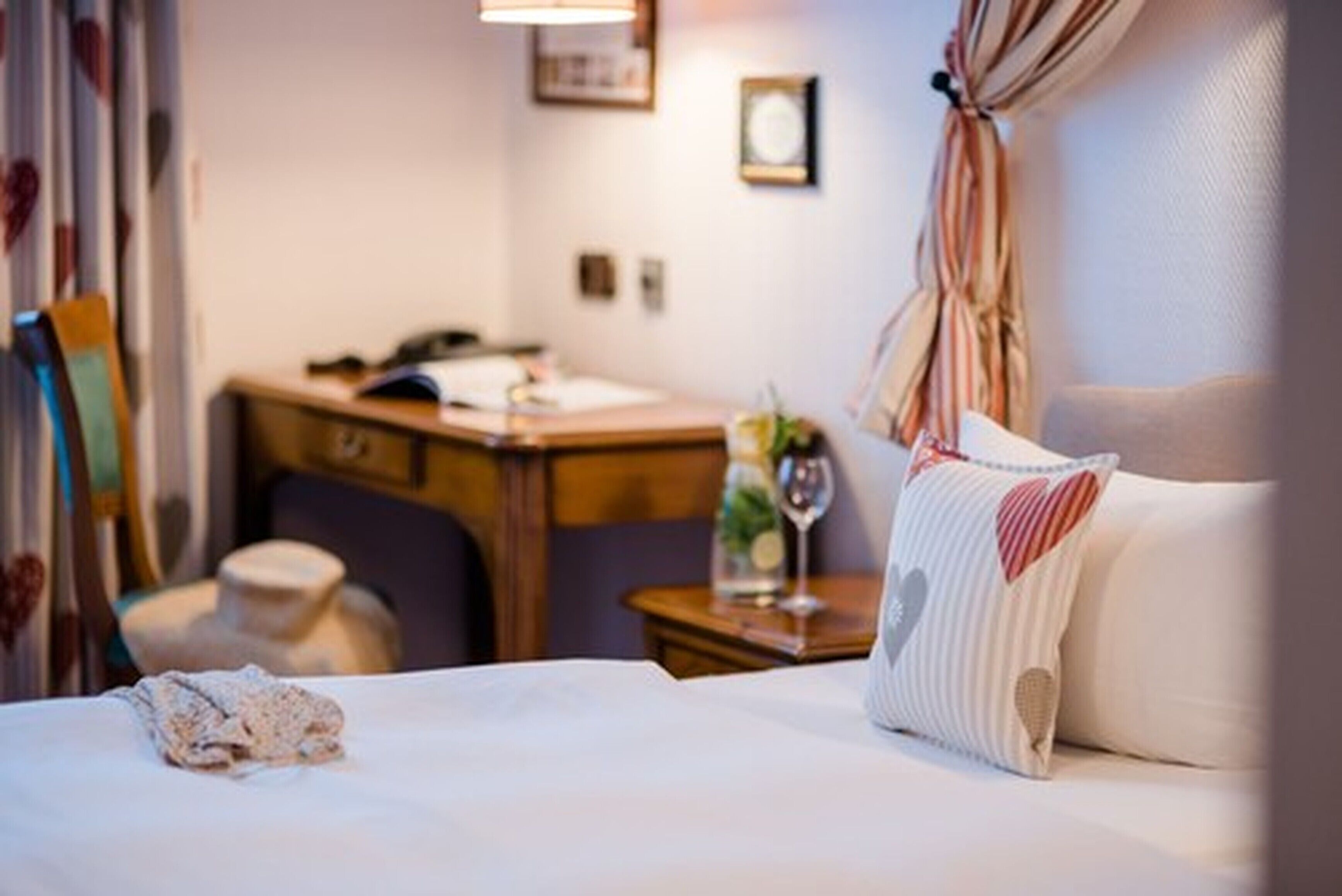 A hotel room with a bed, a desk, a chair, and a small table with a vase and a glass.
