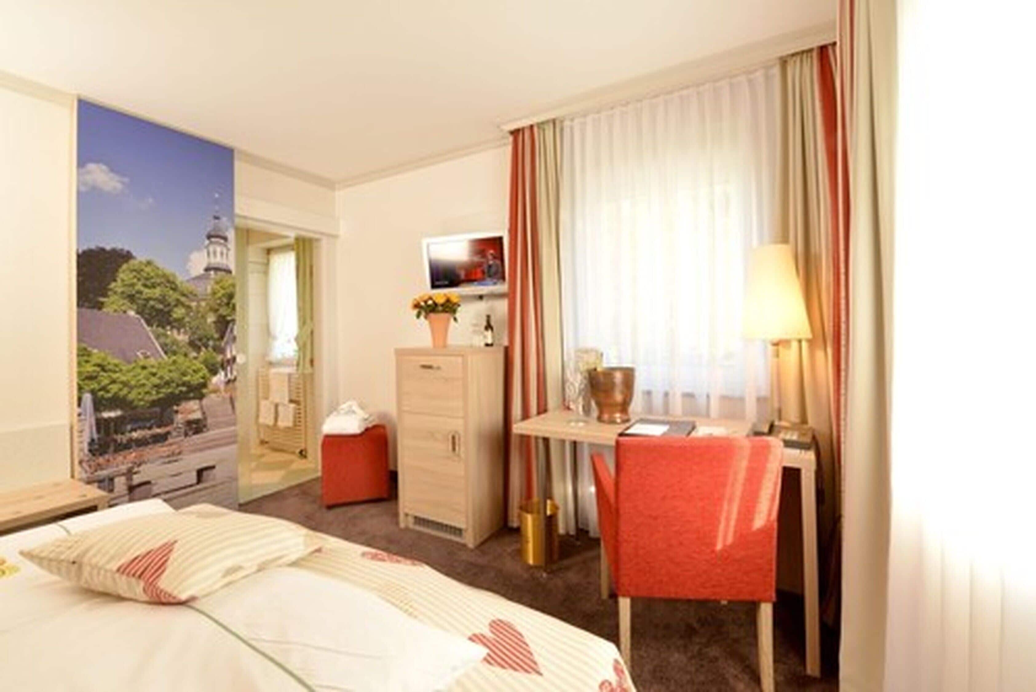 Una habitación de hotel con cama, escritorio con silla, televisión y ventana con cortinas.