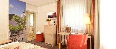 Una habitación de hotel con cama, escritorio con silla, televisión y ventana con cortinas.