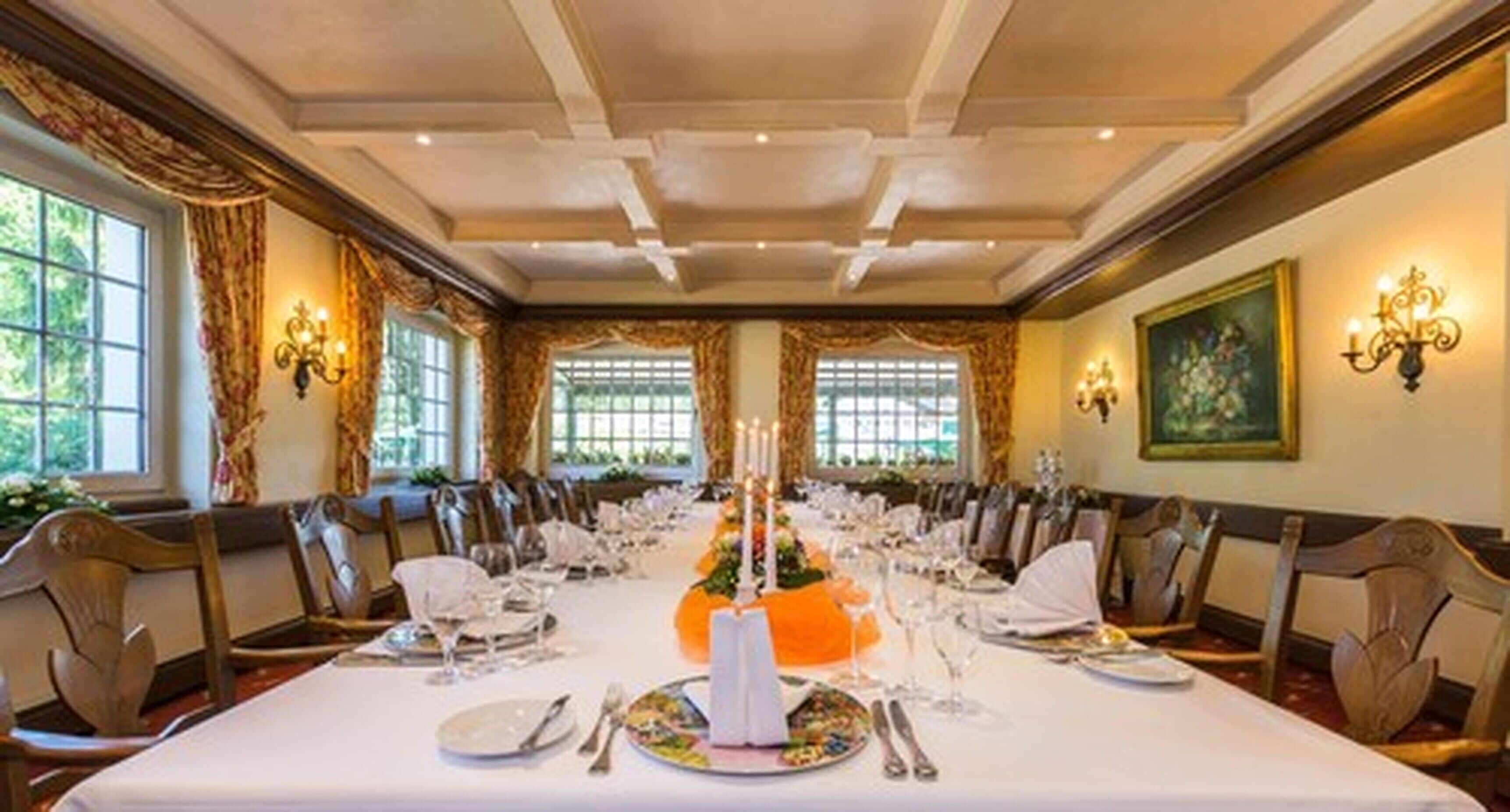 A formal dining room with a long table set for a meal, flanked by chairs, with a chandelier and wall sconces providing lighting.