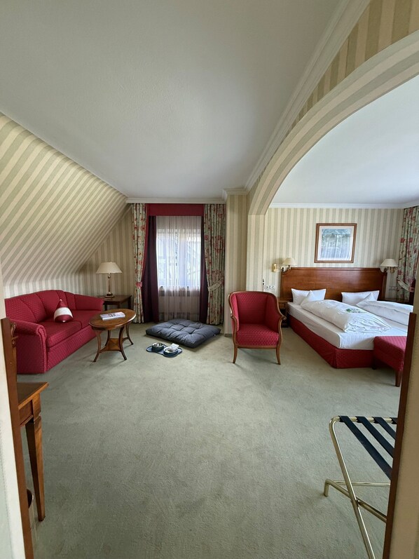 Quarto de hotel com cama, sofá, cadeira, mesa pequena e janela com cortinas.