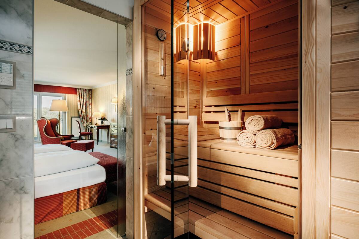 Una habitación de hotel moderna con sauna de madera, baño con paredes de vidrio y una zona de estar acogedora con una butaca roja y un escritorio.
