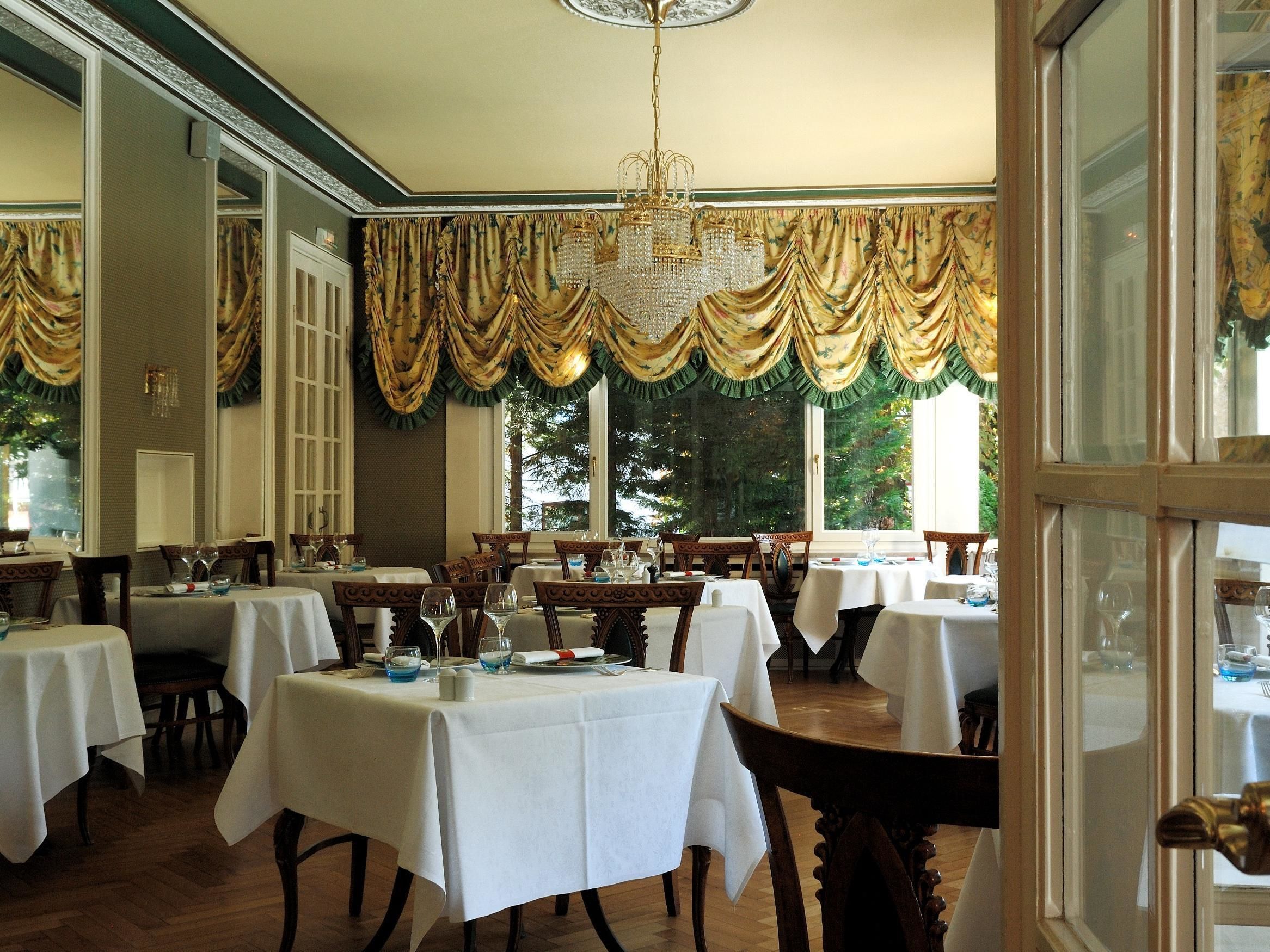 A dining area with multiple tables set for service, large windows with curtains, and a chandelier.