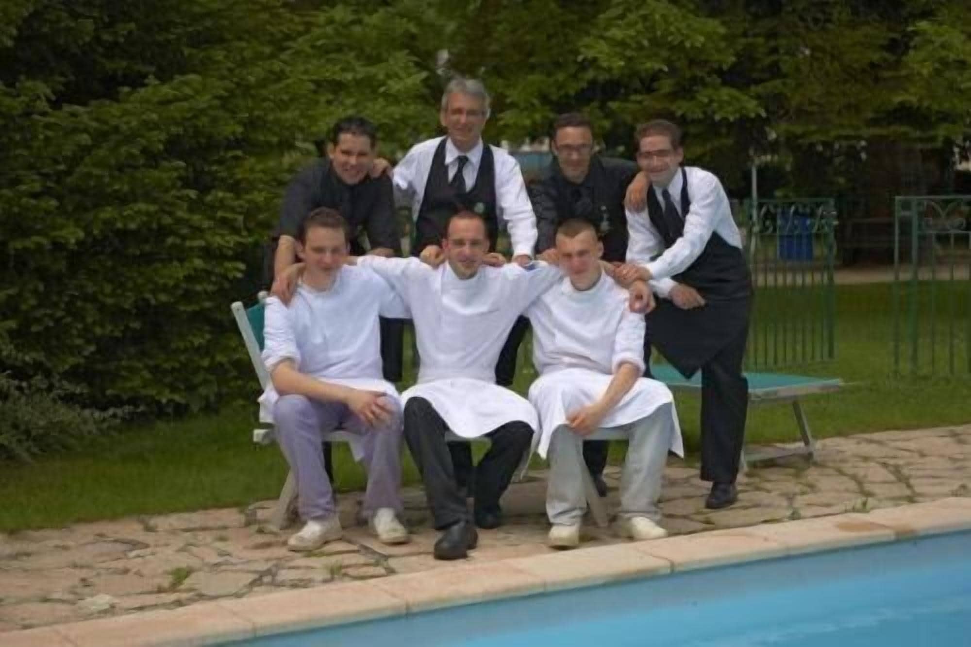 A group of men posing by a poolside with a stone pathway and greenery in the background.
