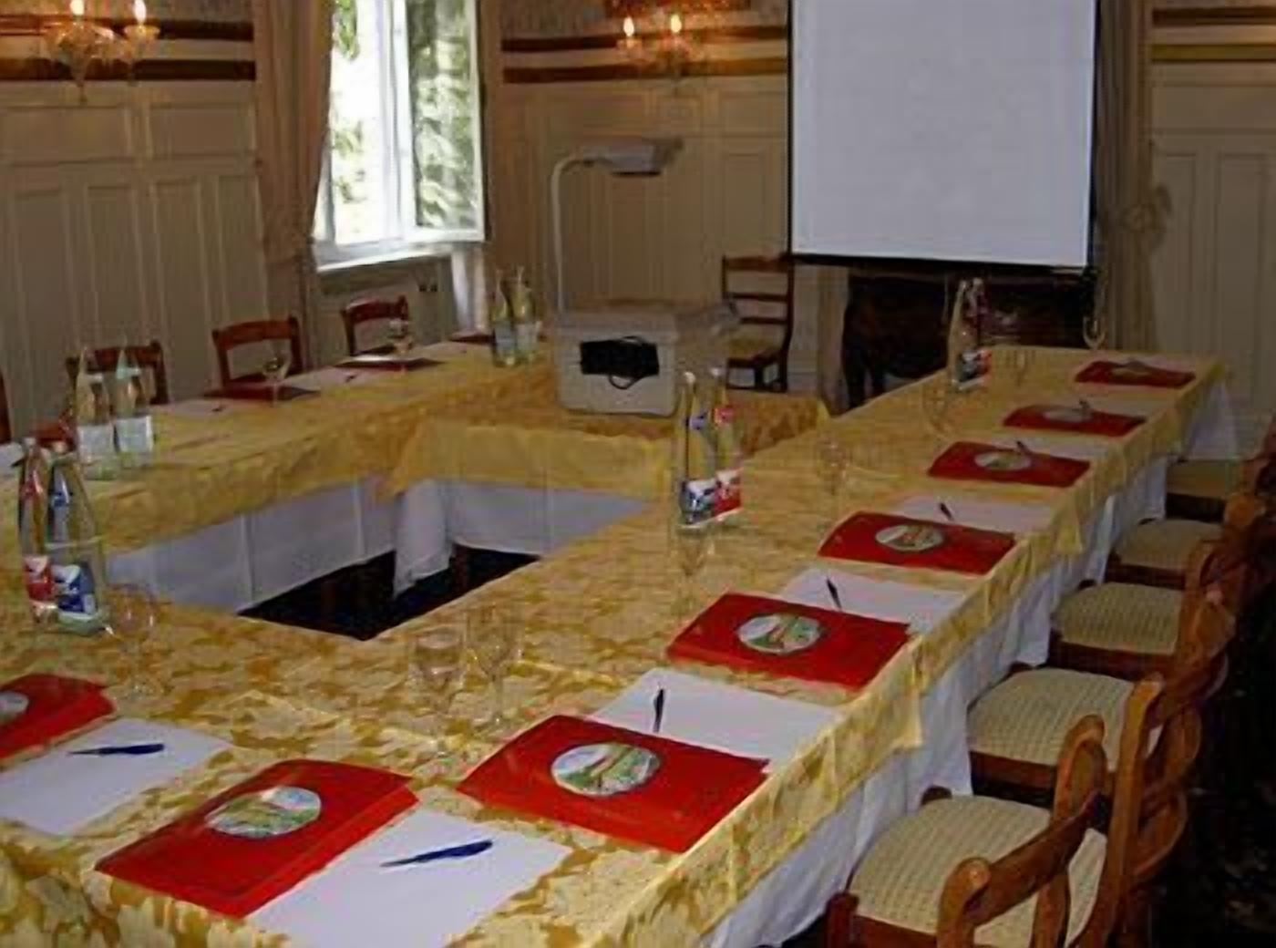 Une salle de conférence avec une longue table préparée pour une réunion, équipée de porte-noms, de bouteilles d’eau et d’un écran de projection.
