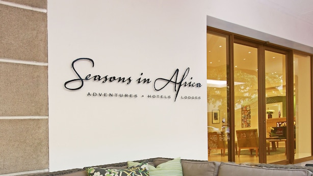 Un espace extérieur moderne avec un canapé et une table basse en verre, une enseigne indiquant « Les saisons en Afrique », et un aperçu de l’intérieur visible à travers une porte vitrée.