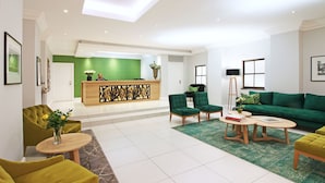 Un hall d’hôtel moderne avec un comptoir d’accueil, des fauteuils et un canapé recouverts d’un tissu vert, et un sol carrelé en marbre.