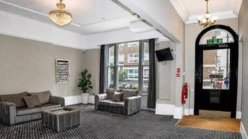 A hotel lobby with a checkered floor, two sofas, a potted plant, a chandelier, and a fire exit door.