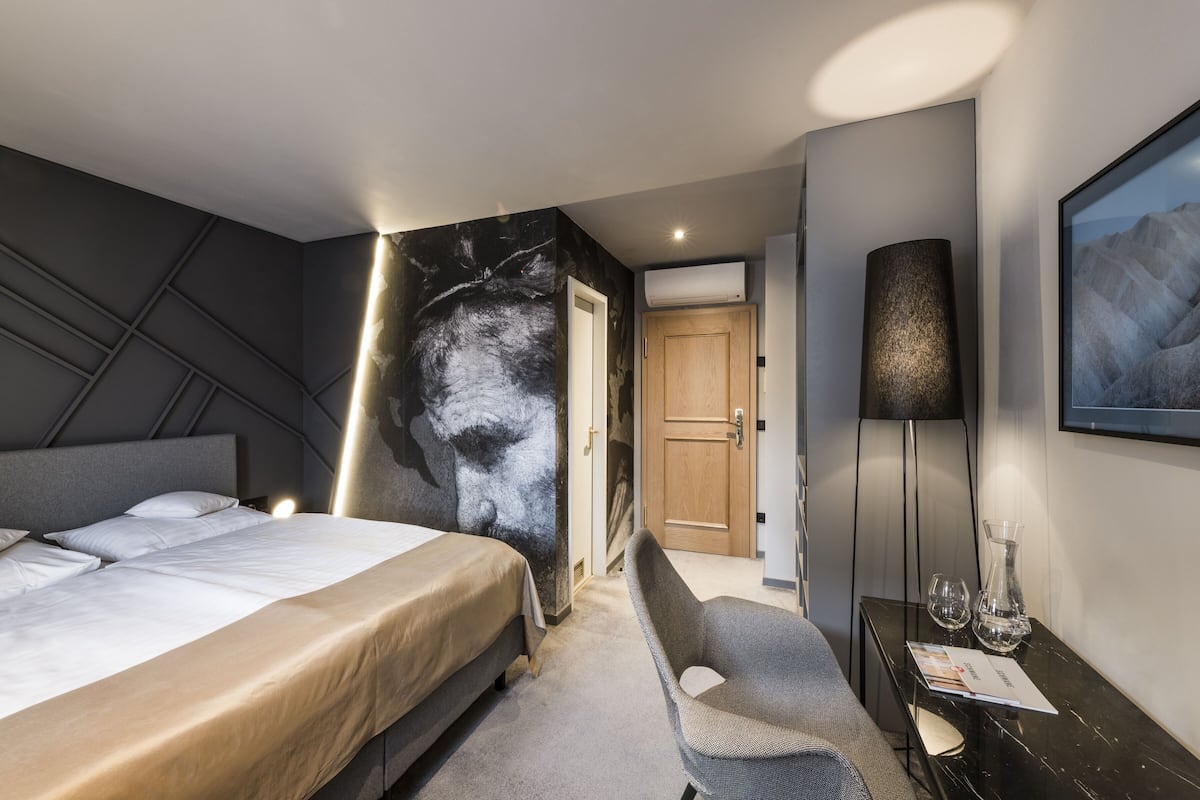 A modern hotel room with a large bed, a seating area, a desk with a lamp, and a large framed picture on the wall.