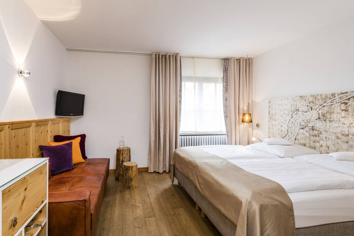 A hotel room with a wooden headboard, a bed with white linens, a wooden bench with cushions, a small table with glasses, and a television mounted on the wall.