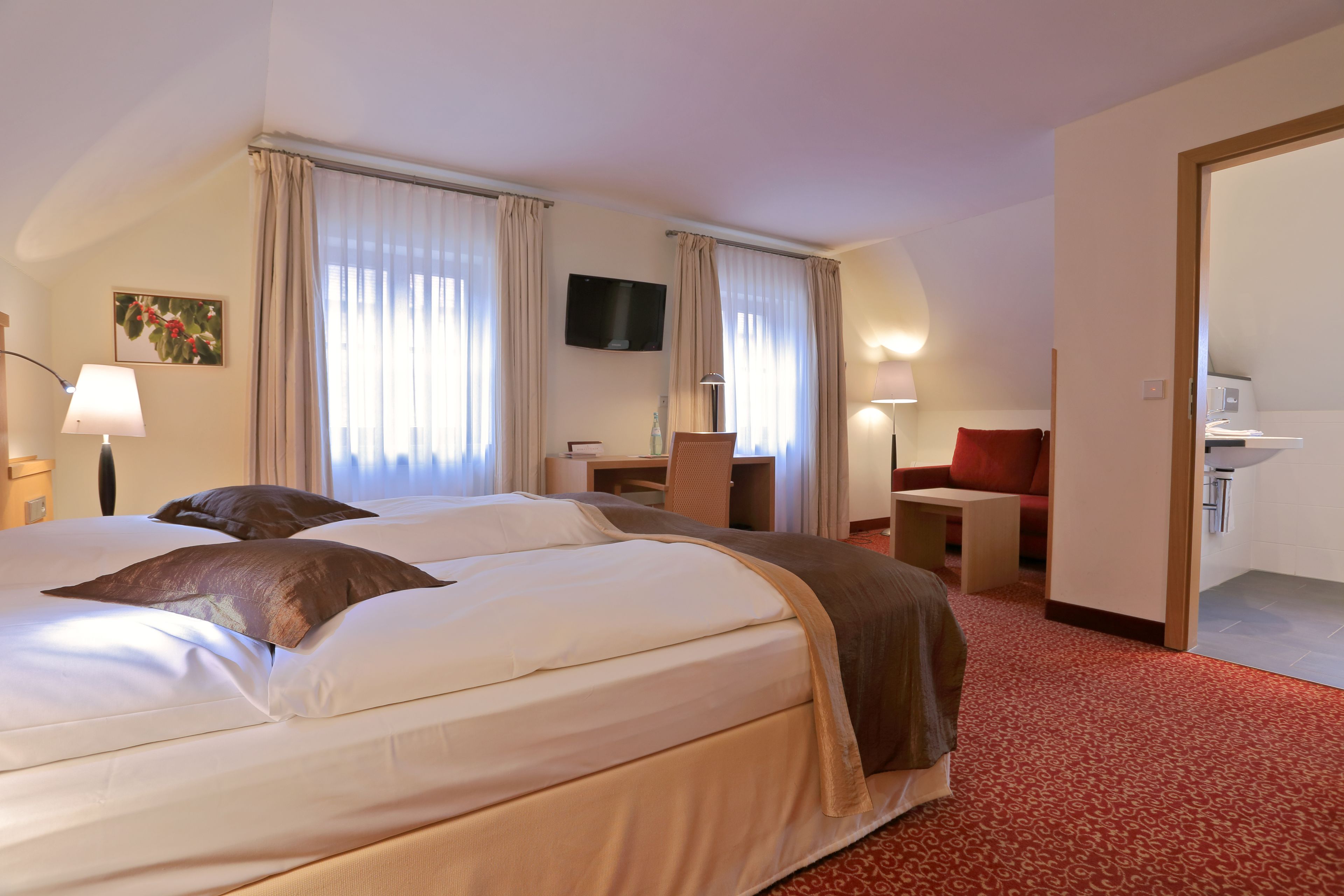 A hotel room with a large bed, a desk, a TV, and a bathroom visible through an open door.