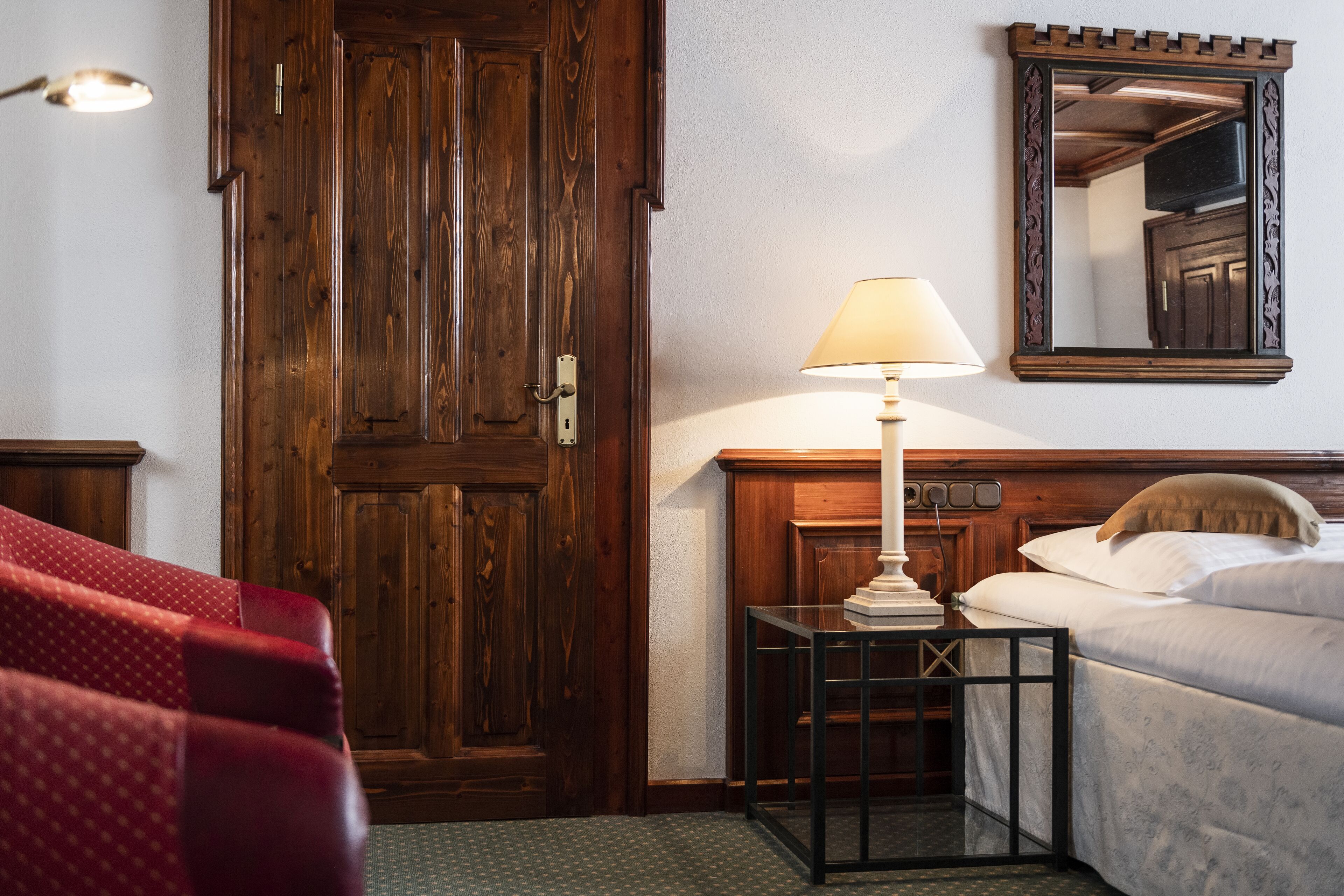 A hotel room with a wooden door, a bedside table with a lamp, a bed, and a red upholstered chair.