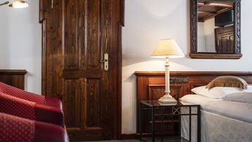 A hotel room with a wooden door, a bedside table with a lamp, a bed, and a red upholstered chair.