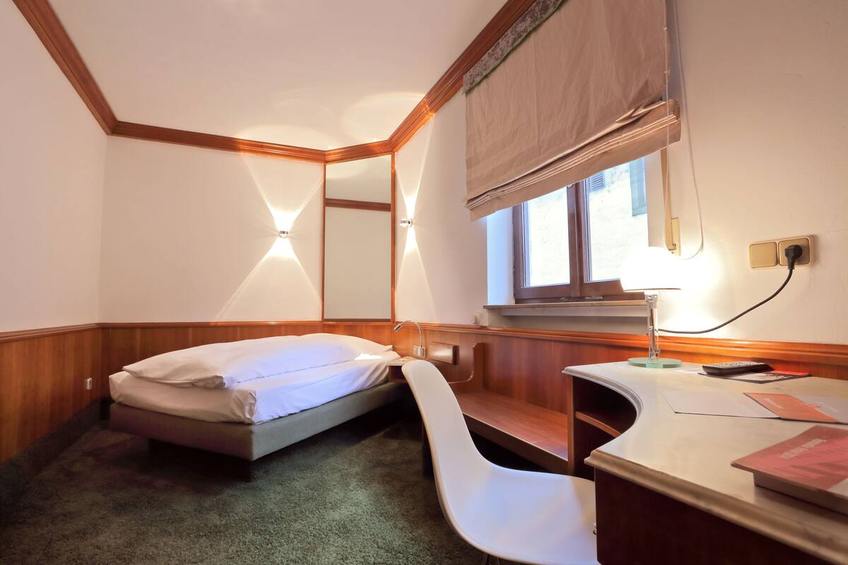 A small, compact room with a single bed, a desk with a chair, and a window with a blind.