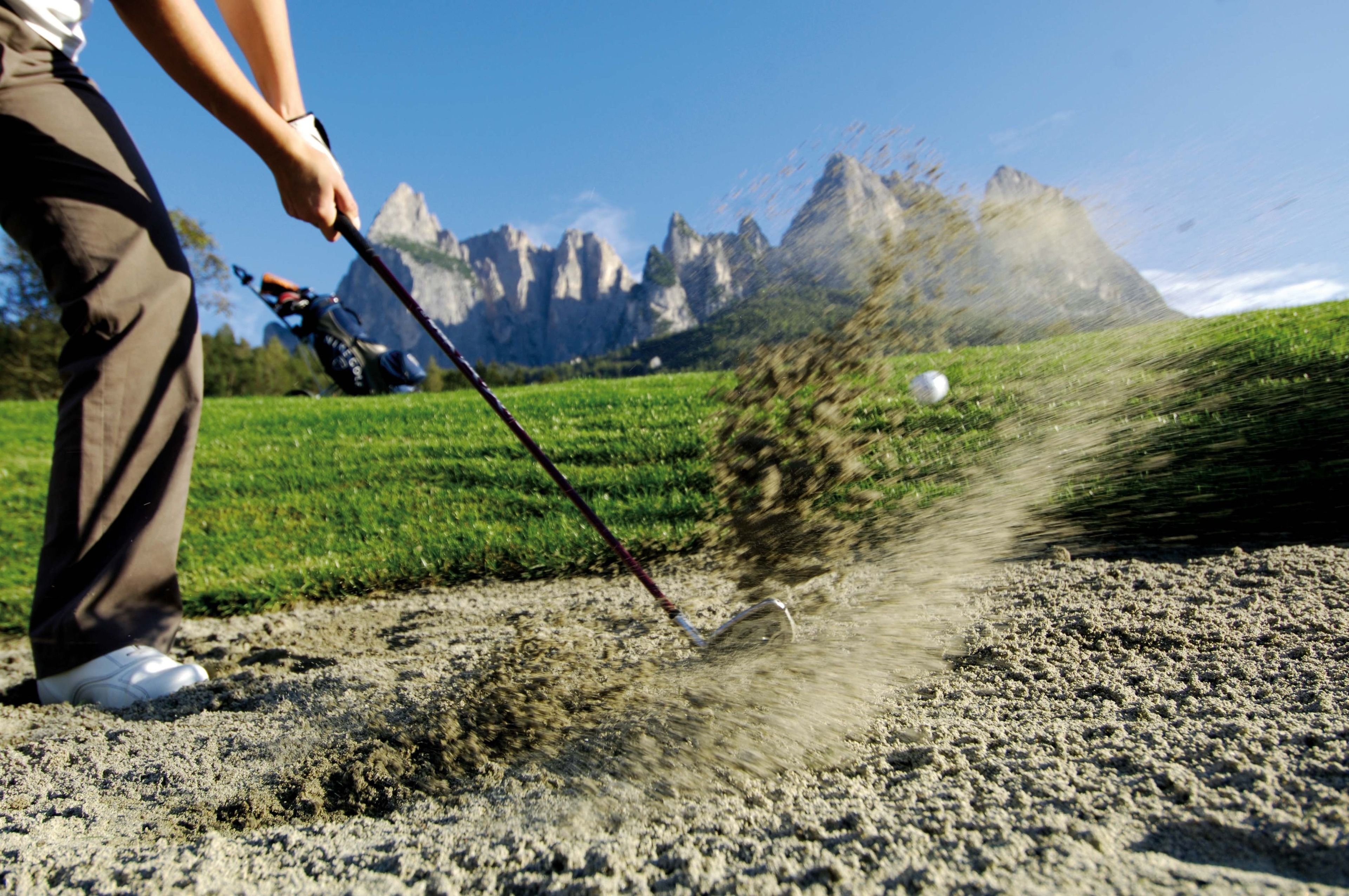 Eine Person schwingt einen Golfschläger in einer sandigen Umgebung mit Bergen im Hintergrund.