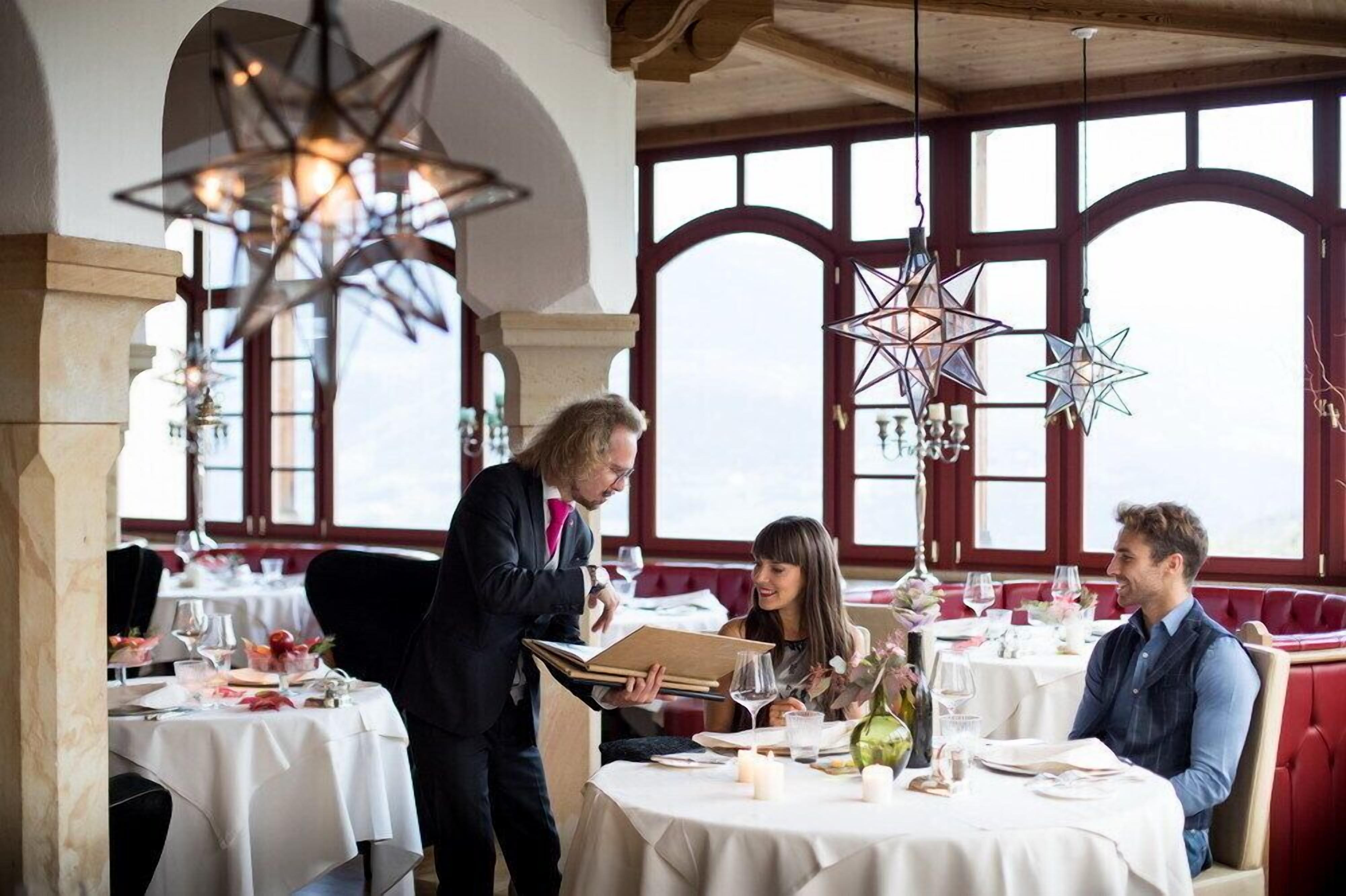 Un restaurante con mesas preparadas para comer, un camarero atendiendo una carta y vistas del cielo a través de amplios ventanales.