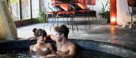 A couple in a heated indoor pool with a view of a dining area and chairs.