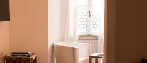 A bedroom with a wooden floor, a white bed, a small table, and a window with curtains.