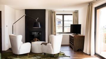 Un salon moderne avec une cheminée, deux fauteuils blancs, une table basse, un téléviseur posé sur un meuble en bois et une grande fenêtre avec des rideaux.