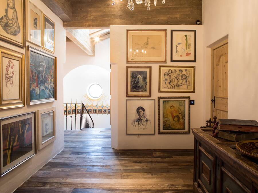 Um corredor com obras de arte emolduradas nas paredes, piso de madeira e uma escada ao fundo.