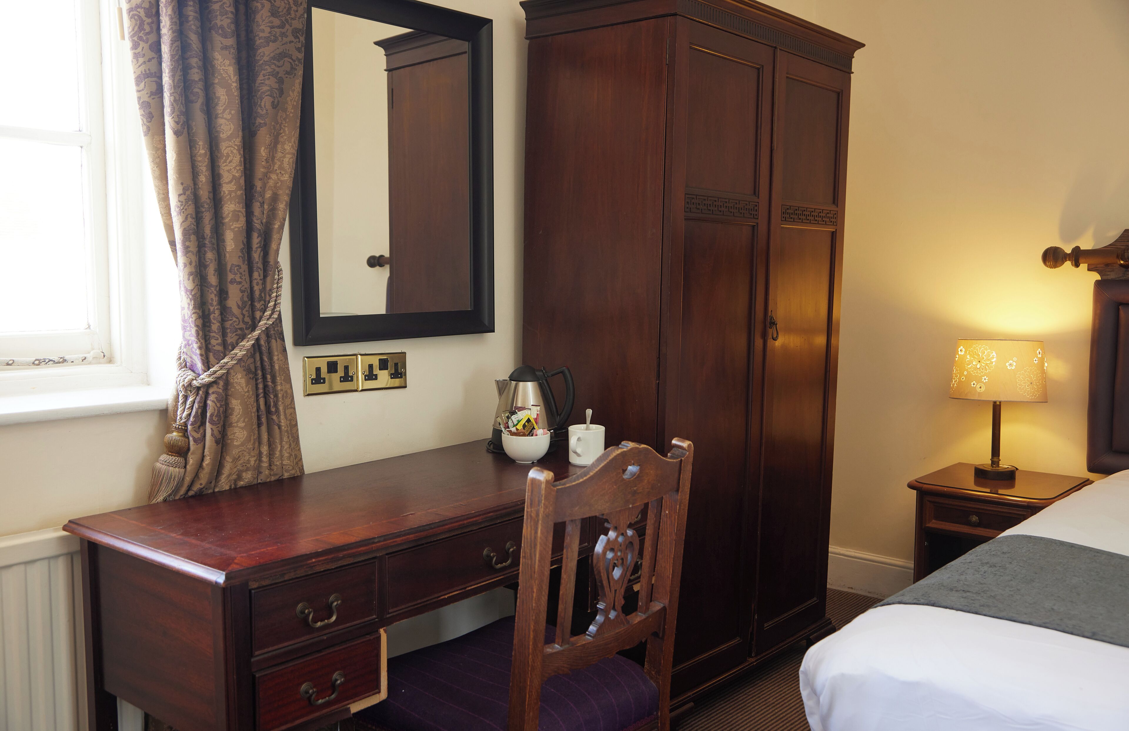 Habitación de hotel con un escritorio de madera, una silla, una cama, un armario y una mesita de noche con lámpara.