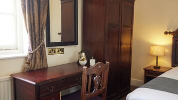 A hotel room with a wooden desk, a chair, a bed, a wardrobe, and a bedside table with a lamp.