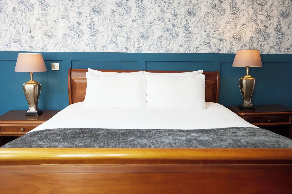 A neatly made bed with white linens, flanked by two bedside tables with lamps, against a wall with floral wallpaper.