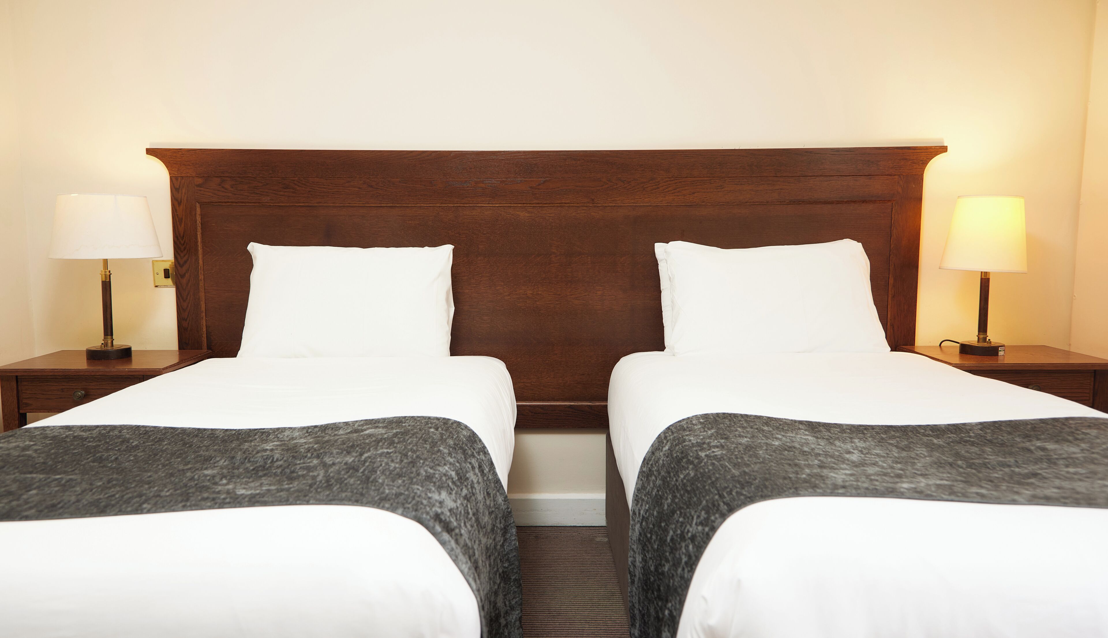 A double room with two beds, each with a wooden headboard, white linens, and a grey throw blanket. There are two bedside tables with lamps, and a wall-mounted light switch.